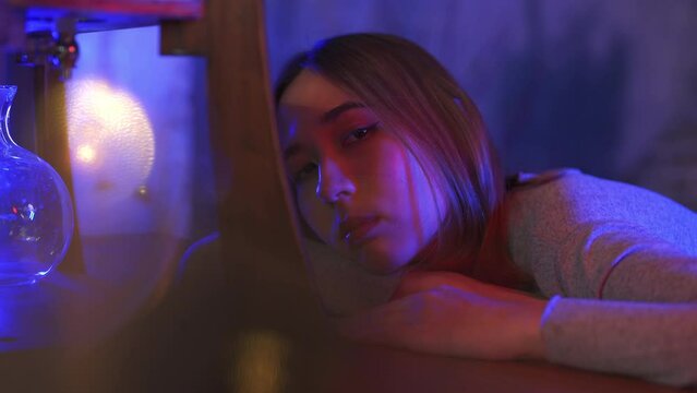 4K Portrait of Loneliness Asian woman sitting on sofa in nightclub with illuminated neon night light with sadness eyes. Beautiful female feeling depressed living alone in multi-colored vibrant lights