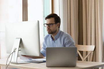 Concentrated young businessman in eyeglasses working on different devices, analyzing information, considering problem solution, creating new online project or doing economic research at home office.