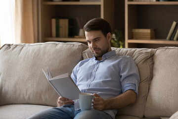 Smiling relaxed young man involved in reading paper book, holding cup of hot tea or black coffee in hands, feeling excited of buying new novel or bestseller, enjoying weekend hobby activity at home.