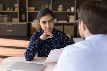 Interested young professional female Indian human resources manager holding paper CV resume in hands, listening skilled male Caucasian job candidate at interview meeting, employment hiring concept.