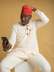 Yoruba Culturally Dressed Business Man Sitting with Phone in Hand and Hand on Cap