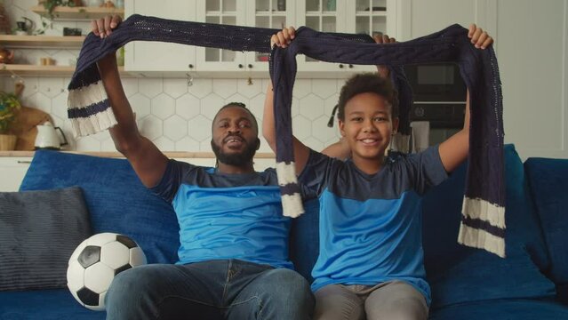 Cheerful handsome black father and happy cute preadolescent son with football scarves watching soccer game on tv, cheering team and chatting slogans while sitting on couch in domestic room.