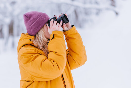 Happy girl photographer in a yellow jacket takes pictures of winter in a snowy park