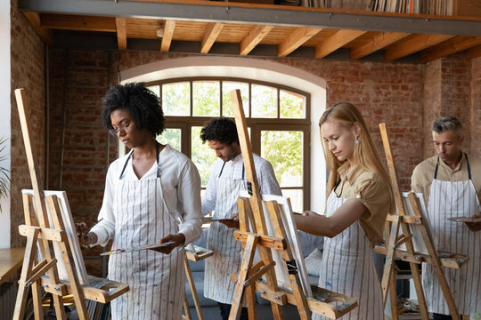 Multiethnic group of artist students drawing at easels on art school class, using paints, palette, easels, canvas, brushes in studio, enjoying craft hobby, creative therapy