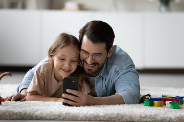 Cute girl lying on carpet with dad watch new vlog on cellphone, use smartphone for leisure fun enjoy cool mobile application smiling resting on floor in playroom. Modern tech, parental control concept