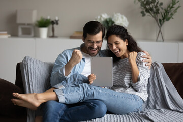Happy overjoyed couple sit on sofa look at digital table screen scream with joy clenched fists...