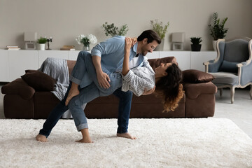 Loving husband dancing tango with lovely wife, standing barefoot on carpet in modern living room with fashionable furniture, full-length view. Wedding dance rehearsal, hobby, love, celebration concept