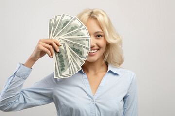 Portrait of happy business lady hiding face behind cash, holding bunch of dollars and smiling over light grey background
