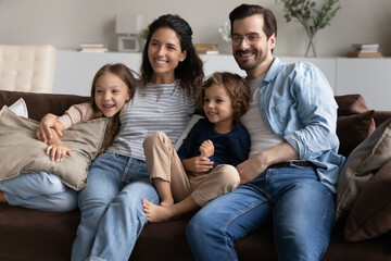 Married young couple and preschool siblings sitting on sofa relaxing together at modern home smiling looking into distance. Adoption and custody, young homeowners family portrait, bank loan concept