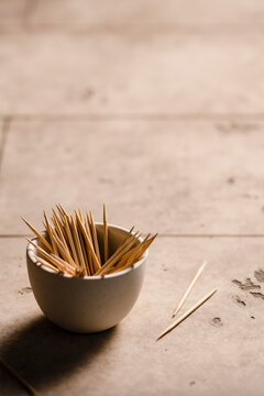 A small bowl of toothpicks on the edge of a table with a few scattered on the table.