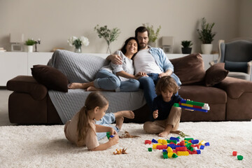 Young couple relaxing on sofa in living room looking at their children play with colorful bricks sitting on warm floor on carpet. Family weekend leisure, modern playthings store ad, pastime concept