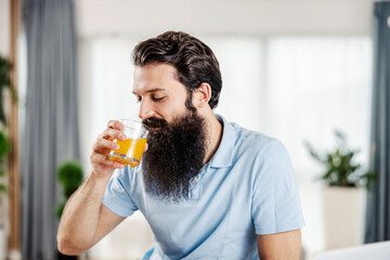 Fototapeta premium A bearded man drinking fresh orange juice while sitting at his cozy home in the morning.