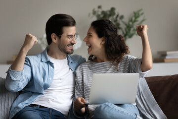 Couple sit on sofa with laptop clenched fists make yes gesture celebrate great news. Got bank loan acceptance, achievement, success, online lottery, bet victory, winners family, candid emotion concept