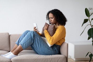 Depressed young black lady reading sad news or message on smartphone, sitting on couch and hugging pillow at home