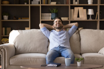 Laughing cheerful young male homeowner sitting on comfortable sofa, breathing fresh air, enjoying carefree pleasant weekend leisure time alone in modern living room, meditating or resting indoors.