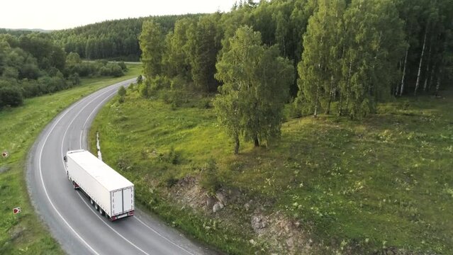 Aerial drone view of a truck car driving on a bending road among green trees. Scene. White long cargo car on the empty road near summer forest.