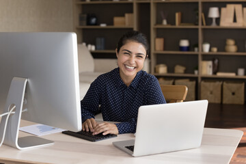 Portrait of happy successful beautiful young Indian businesswoman employee sitting at workplace with laptop and monitor, using different gadgets for work in modern office room, career concept.