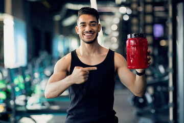 Smiling Sporty Arab Guy Advertising Fitness Supplements While Posing At Gym Interior