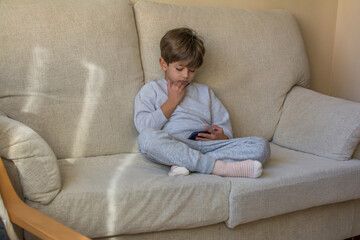 6 Years Old Boy In Casual Clothes Sitting On Sofa And Playing Video Games On Smartphone On Weekend At Home. Children and Technology