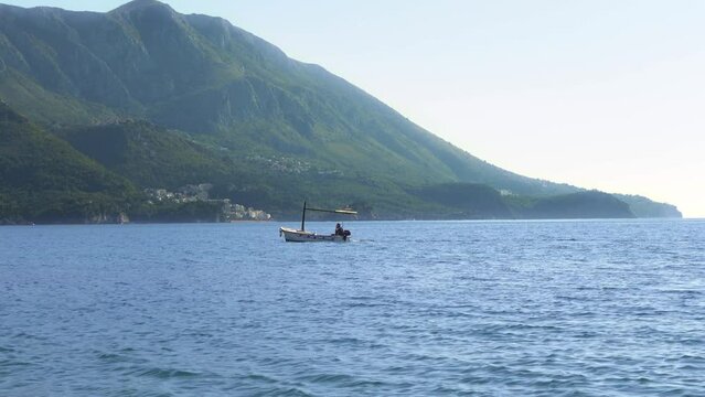 Small fishing boat sails in Adriatic sea against coastal village at forestry mountain foot ON sunny day Budva, Montenegro