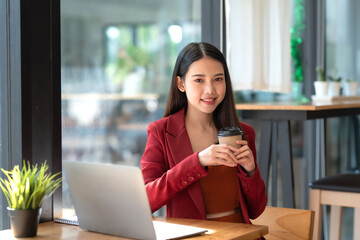 Confident beautiful young Asian businesswoman in a red suit sitting holding coffee with laptop computer in café. Looking at camera.