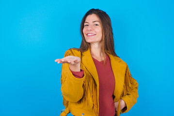 young brunette woman wearing yellow fringed jacket over blue background smiling friendly offering something with open hand or handshake as greeting and welcoming. Successful business.