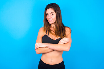 Waist up shot of  self confident Young beautiful sportswoman doing sport wearing sportswear over blue background has broad smile, crosses arms, happy to meet with colleagues.