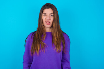 Fototapeta premium Portrait of dissatisfied young beautiful caucasian woman wearing purple sweater against blue smirks face, purses lips and looks with annoyance at camera, discontent hearing something unpleasant