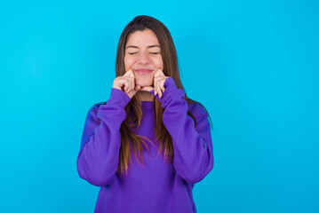 Pleased young beautiful caucasian woman wearing purple sweater against blue background with closed eyes keeps hands near cheeks and smiles tenderly imagines something very pleasant