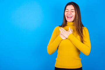 young beautiful caucasian woman wearing yellow turtleneck sweater against blue background expresses happines, laughs pleasantly, keeps hands on heart