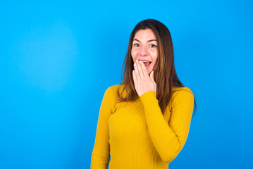 young beautiful caucasian woman wearing yellow turtleneck sweater against blue background covers mouth and looks with wonder at camera, cannot believe unexpected rumors.