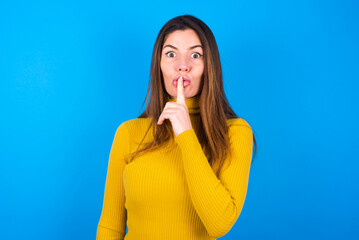 Surprised young beautiful caucasian woman wearing yellow turtleneck sweater against blue background makes silence gesture, keeps finger over lips and looks mysterious at camera