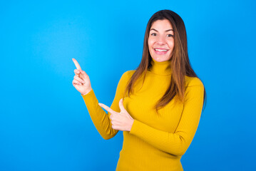 young beautiful caucasian woman wearing yellow turtleneck sweater against blue background points at copy space indicates for advertising gives right direction