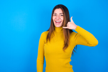 Fototapeta premium young beautiful caucasian woman wearing yellow turtleneck sweater against blue background makes phone gesture, says call me back again, has glad expression.