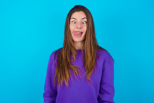 Funny young beautiful caucasian woman wearing purple sweater against blue background makes grimace and crosses eyes plays fool has fun alone sticks out tongue.