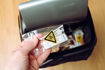 Safe handling and management of rapid antigen COVID-19 testing waste concept. Person hand throw away pieces of antigen test kit sealed in plastic bag, biohazard symbol. Home use.