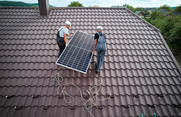 Men technicians lifting photovoltaic solar moduls on roof of house. Electricians in helmets...