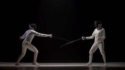 Two female athletes train on foil at fencing tournament. Young female swordsmen in white uniforms and fencing masks fight duel against black background. Fencing sports school. Slow motion. - Powered by Adobe