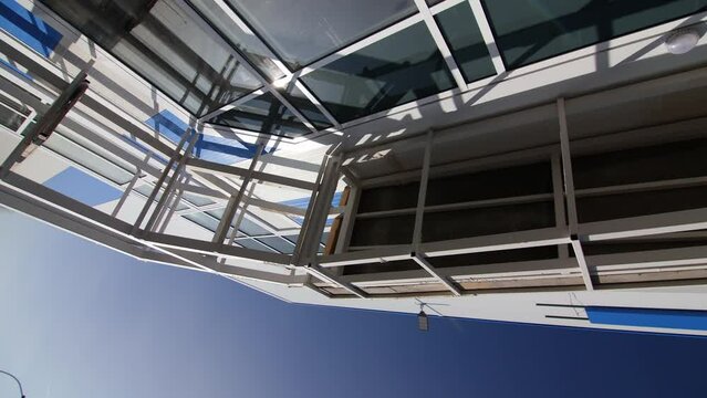 White framework on facade of contemporary building with large dark window reflecting sun dick under blue sky camera rolls closeup