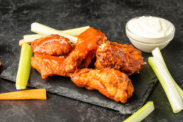 Fried buffalo wings on dark slate in sauce and served with fresh vegetables.