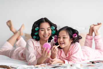 Cheerful millennial japanese lady with curlers hold mirror for teen girl, apply lipstick on lips, on bed with cosmetics