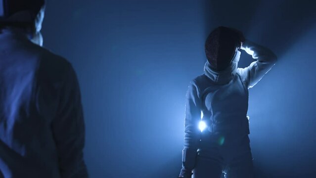 View from behind on two athletes fencers competing in tournament. Women in fencing masks attack each other, striking with epee. Dark studio with backlit blue light and smoke. Slow motion. Close up.