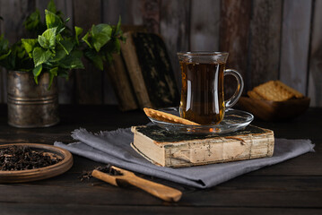 Tea in a glass cup on an old book on a light gray napkin. Mint leaves in a tin mug and cookies in a...