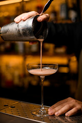 bartender professionally pours brown cocktail from shaker into glass standing on metal bar counter.