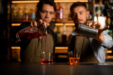 Two glasses stands on bar and two bartenders fills them with alcohol drinks