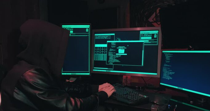 Hacker in a hood sits in front of computer screens and hacks databases. The concept of cyberterrorism, server hacking and cyberattacks. The room is dark, a person is typing code into the command line