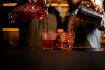 Two bartenders pouring red and brown alcoholic drinks from a shaker and a jug