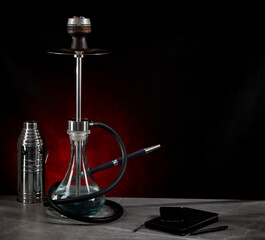 Beautiful modern hookah, stands on the table, surrounded by a metal protective cap and a diary, on a dark red background. Copy space.