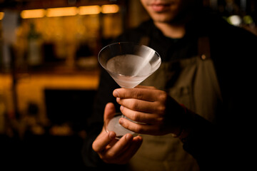 close up photo of bartender in black t-shirt and brown apron holding cooled empty martini glass