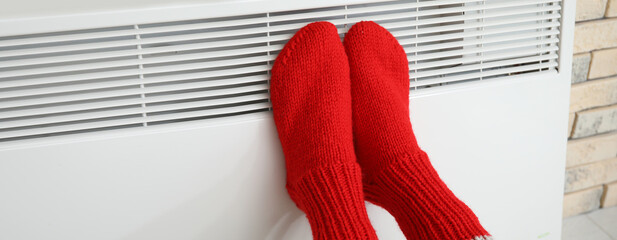 Woman in knitted socks warming her feet at radiator at home. Concept of heating season